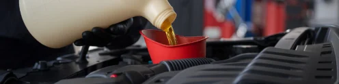 A person wearing black gloves pours engine oil from a large container into a red funnel placed in a car engine bay.